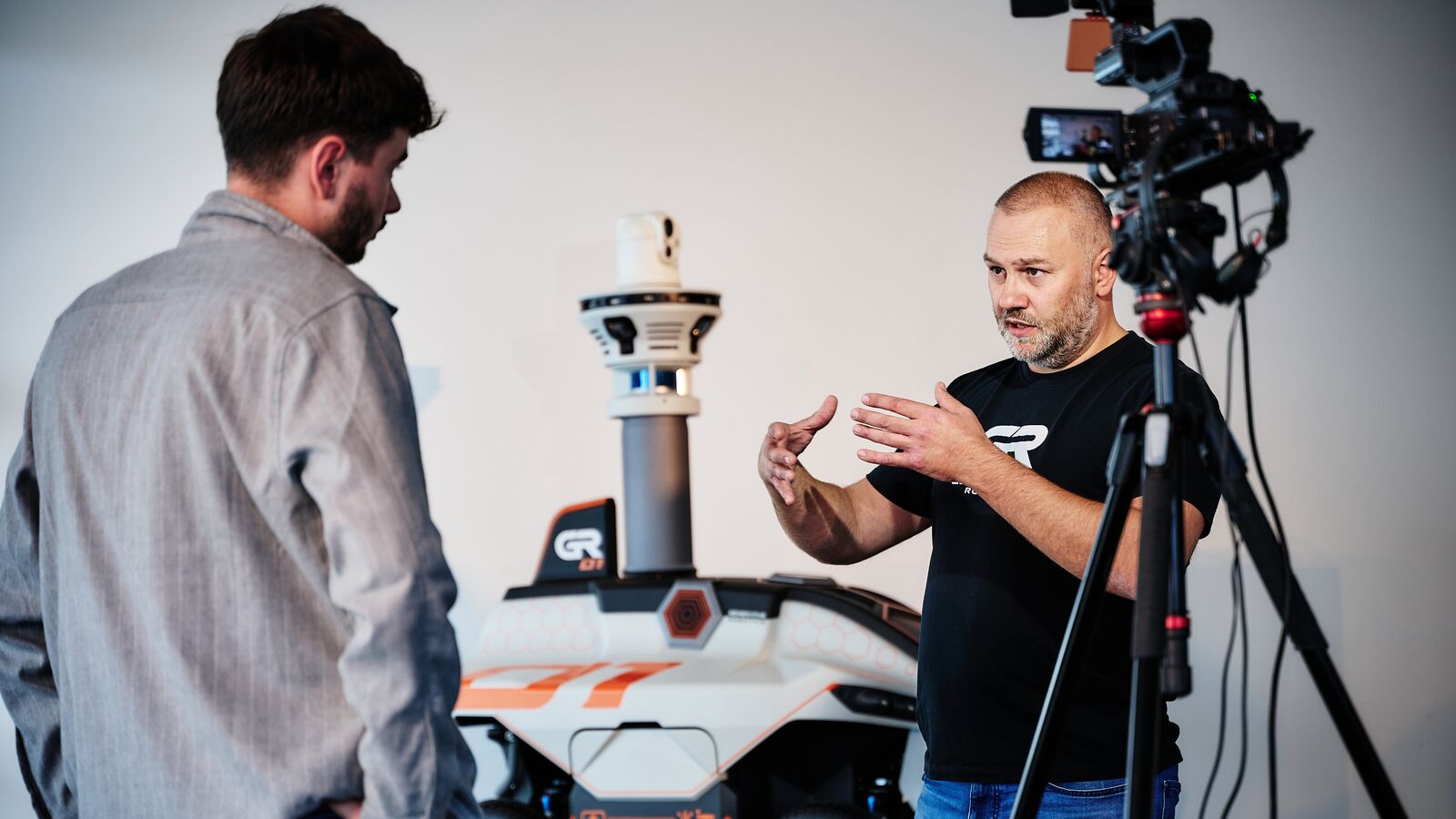 Ein Mann im GENERATIVE-Robotics-Shirt gibt neben dem GR-01 ein Kamerainterview, vor ihm eine Fernsehkamera auf einem Stativ